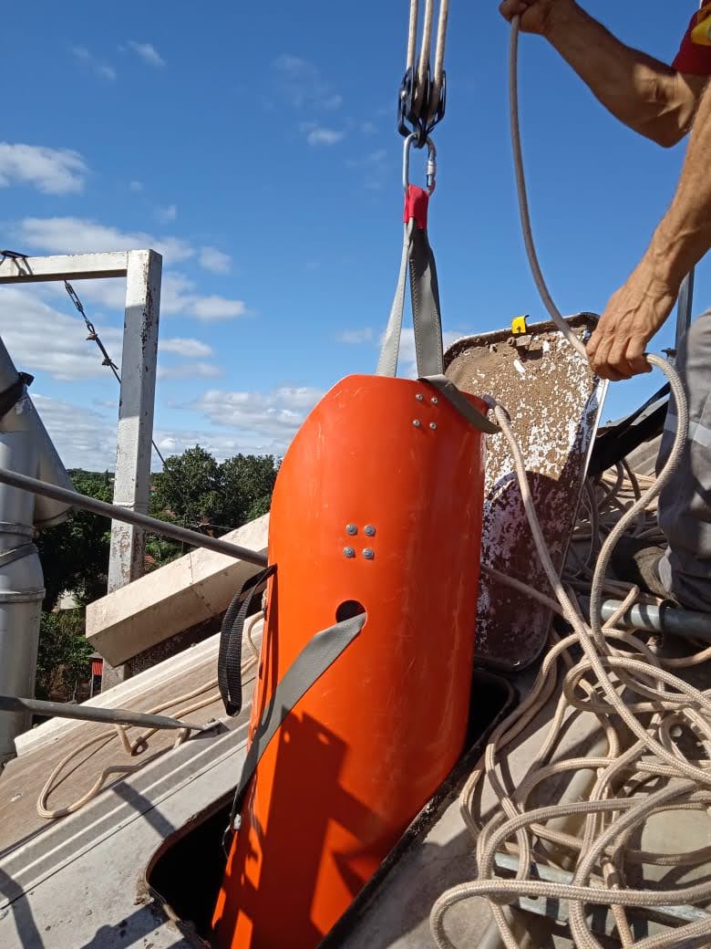 Três engenheiros com capacetes brancos inspecionando uma unidade de grãos com silos ao fundo em um dia ensolarado.